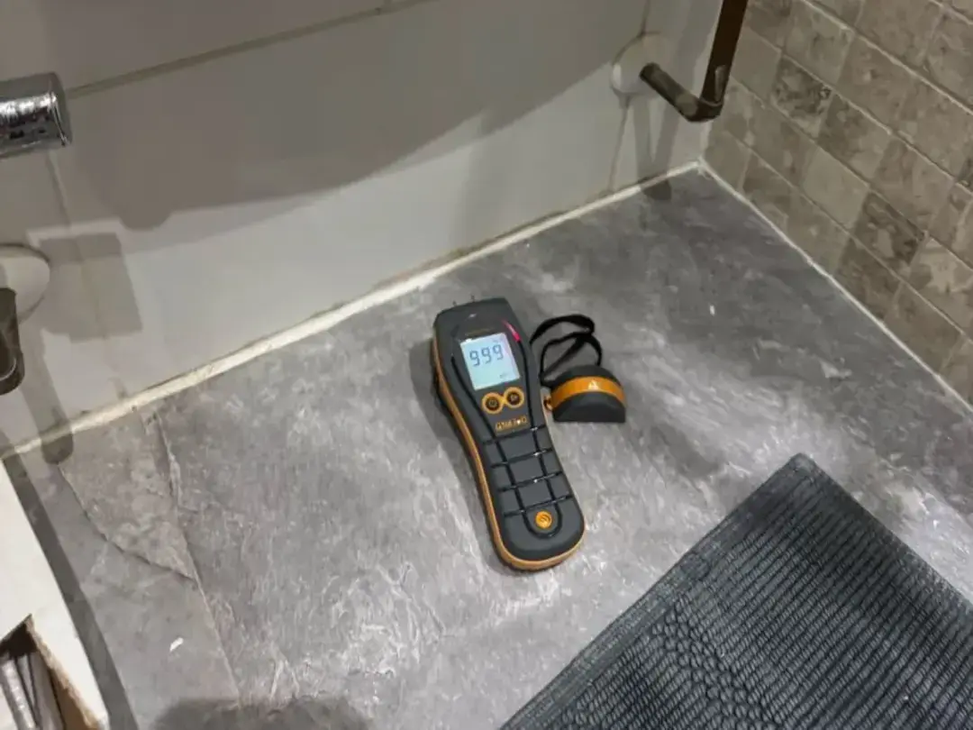 A moisture meter resting on a tiled bathroom floor beside a towel rail
